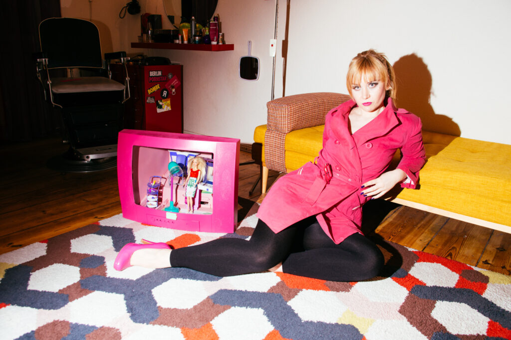 "Young woman in a pink trench coat sits in front of a sofa. Next to her is a pink TV with a miniature Barbie hairdresser salon inside."
